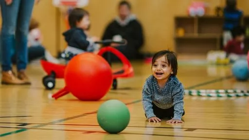 A smiling toddler boy crawls toward a ball on a YMCA gymnasium floor