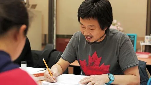 A man wearing a t-shirt with a maple leaf on the front writes in a workbook while speaking with a woman sitting across the table from him
