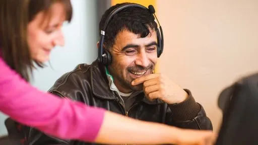 A smiling man wearing a headset looks at a computer screen where a woman standing next to him is pointing