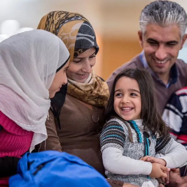 A smiling family. A young girl sits in her mother's lap while speaking to a young adult female, while her father looks on from the background.