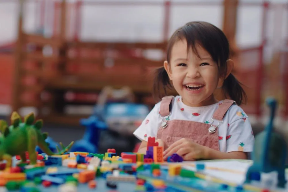 A preschool-aged girl plays with toy bricks on a table outside at a child care centre