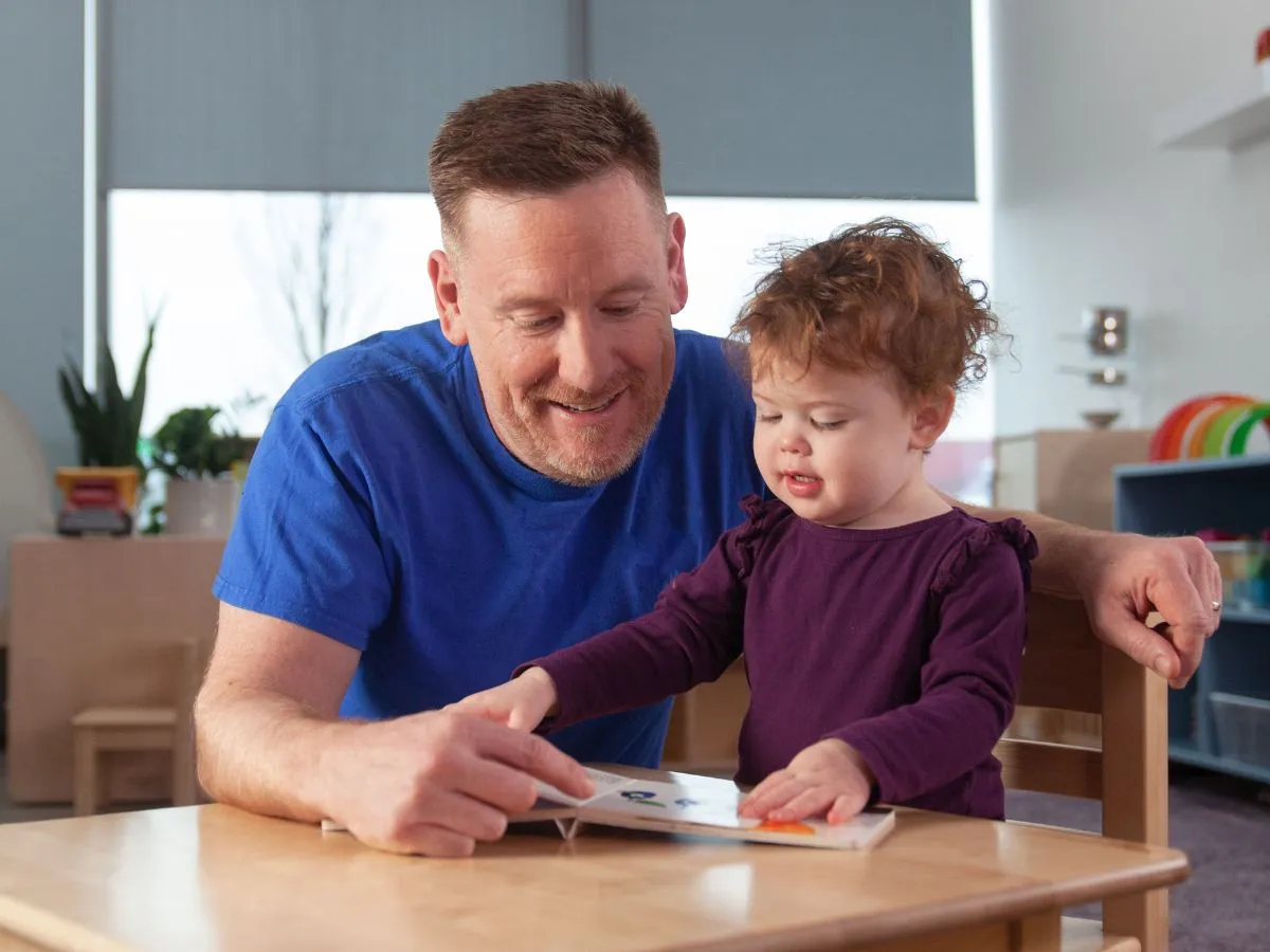 A man and a toddler sit at a table reading a book together.