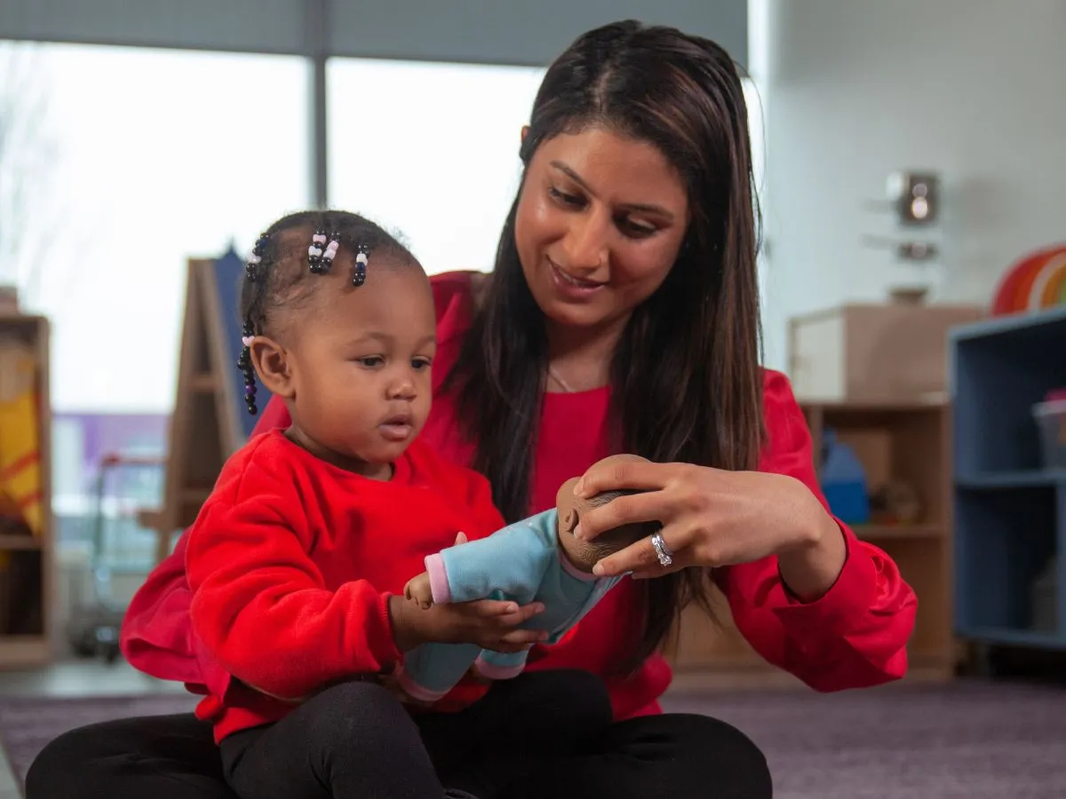 A woman sits on the floor with a toddler in her lap, they are both holding a baby doll. 