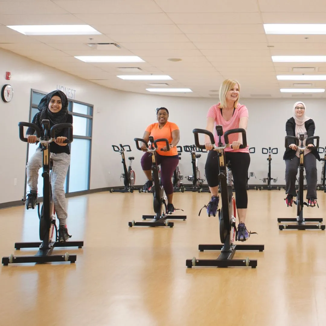 Four women smile while riding stationary bikes in a YMCA fitness studio