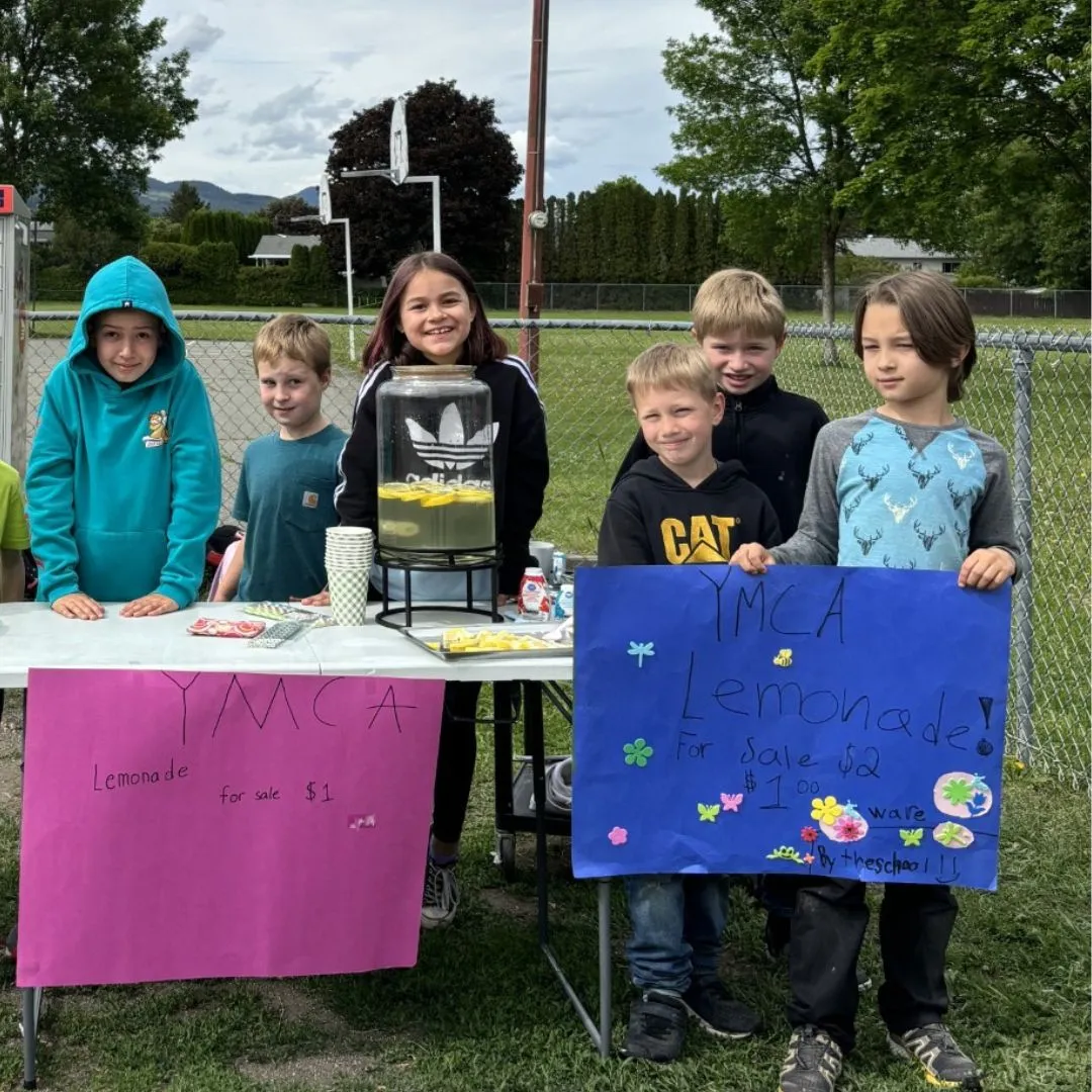 A group of children at a lemonade stand hold signs saying they are raising funds for the YMCA