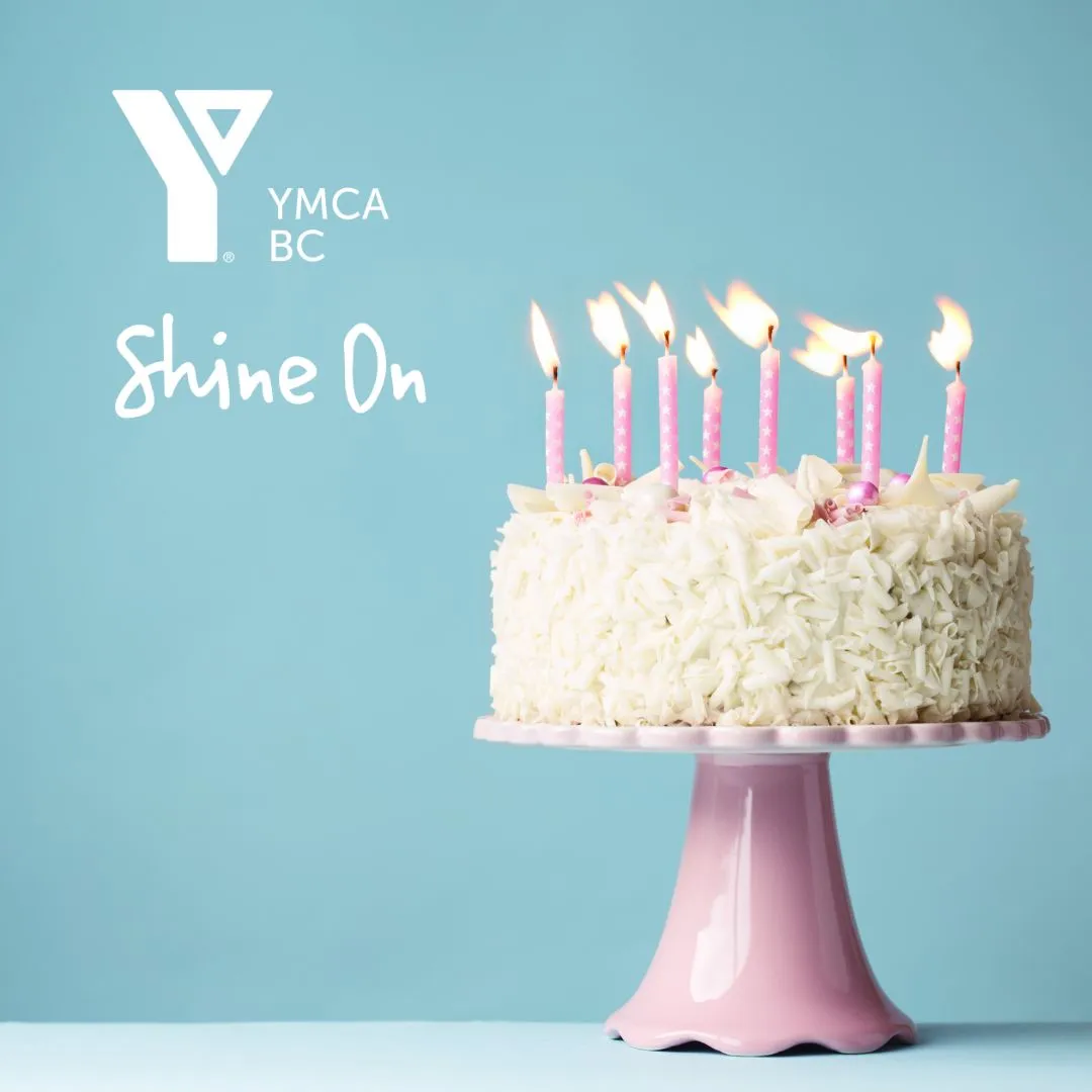 A birthday cake with candles lit next to a YMCA BC logo