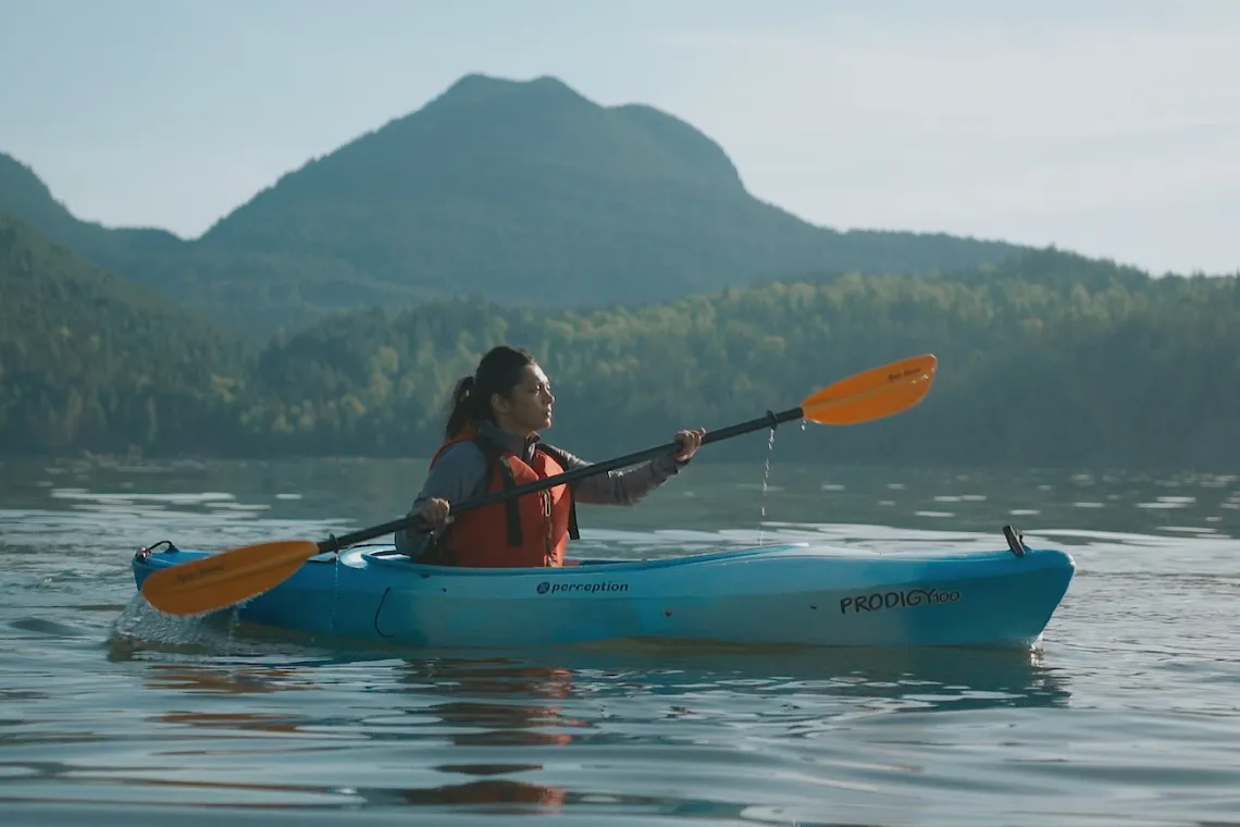 A young adult woman paddles a kayak in the water near YMCA Camp Elphinstone with a mountain backdrop