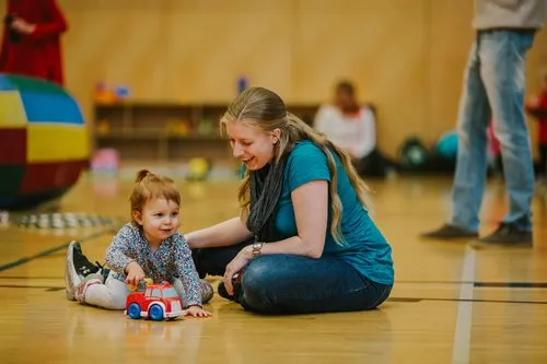 Smiling mother sits on the floor with her toddler daughter, who plays with a truck