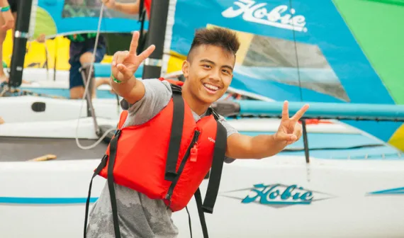 A teen male wearing a lifejacket gives a peace sign from the sailing dock at YMCA Camp Elphinstone