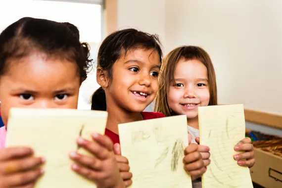 Three young girls hold up what they've drawn and written on paper