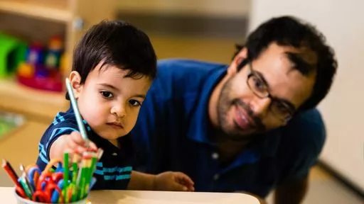 A young boy draws on paper while a smiling man sitting on the floor watches