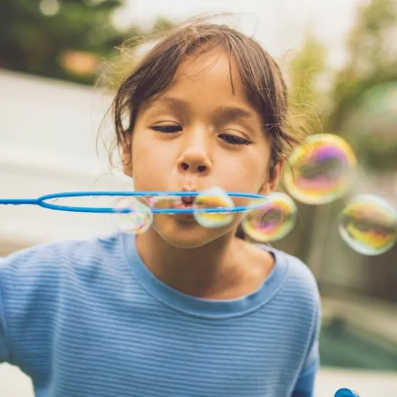 A young girl blows bubbles outside towards the camera