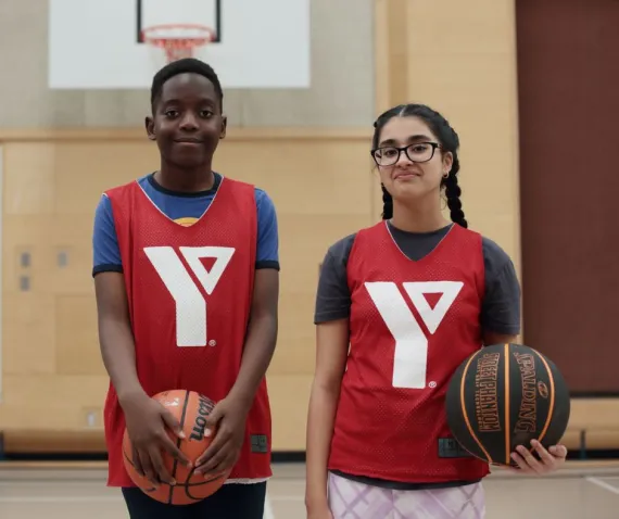 A boy and a girl, both preteen aged, each hold a basketball while wearing YMCA jerseys in a gymnasium
