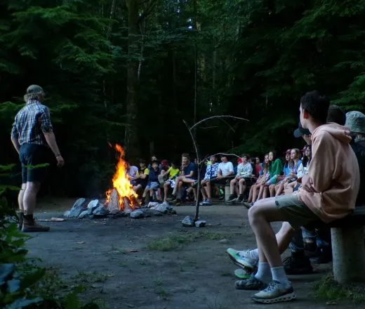 Youth campers are seated around a campfire, listening to a counsellor who is standing near the fire.
