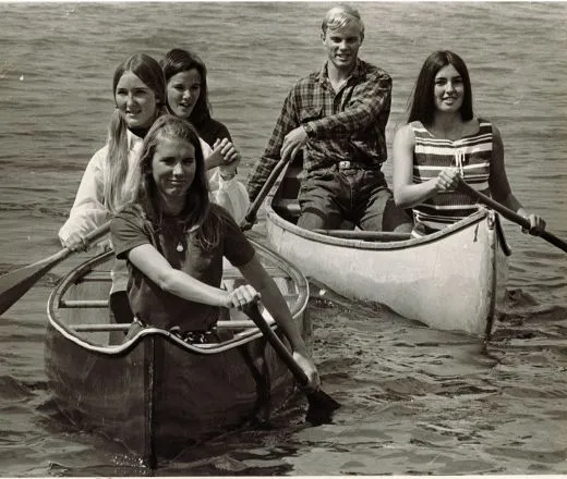 In a black-and-white photo from the 1970s, four teenage girls and one boy paddle two canoes toward shore.