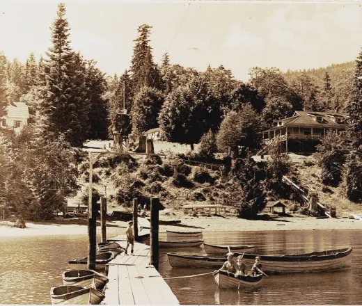 A decades-old photo in sepia tones taken from YMCA Camp Elphinstone's dock, looking back towards the camp shore. Three boys are in a row boat near the dock.