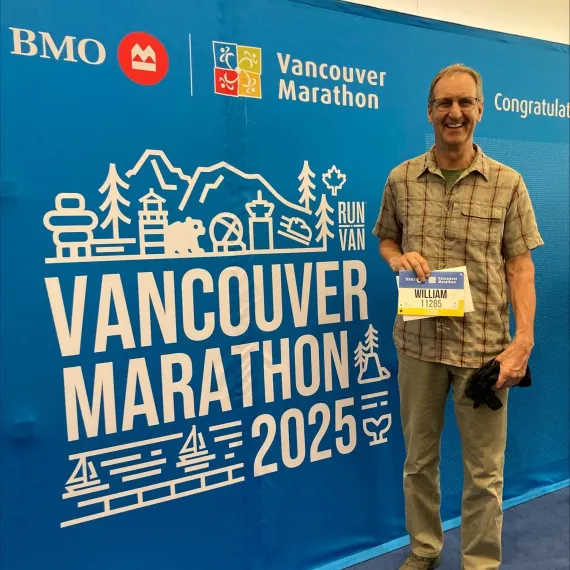 William "Bud" Bruintjes smiles while holding his race bib next to a large sign for the 2025 BMO Vancouver Marathon