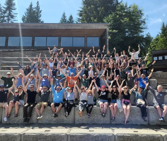 About 100 people sitting outside the dining hall at YMCA Camp Elphinstone hold their arms out in the 'Y' shape