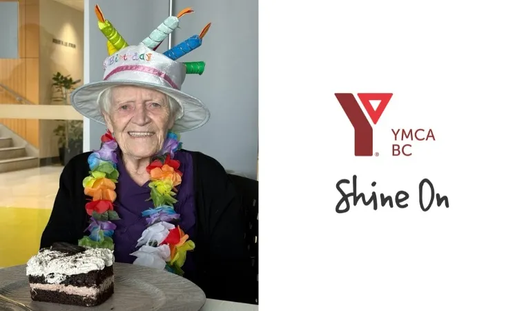 A senior lady smiles with a piece of cake on the table in front of her. She is wearing a floral lei and birthday cake-shaped hat.