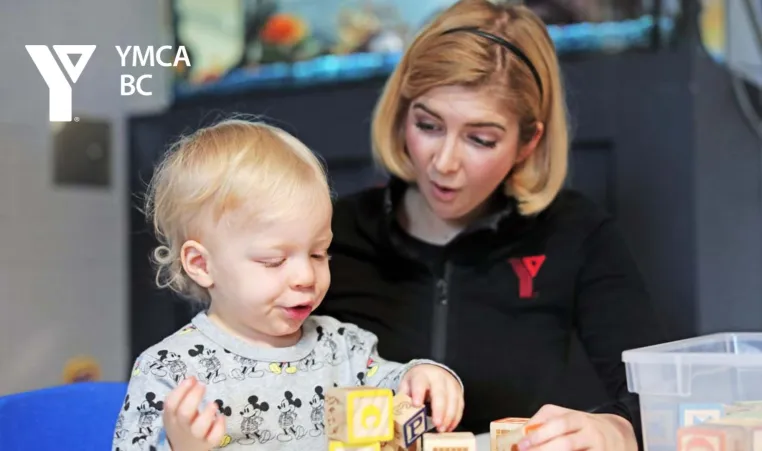 A YMCA child care staff team member sits with and reacts to a young child playing with blocks