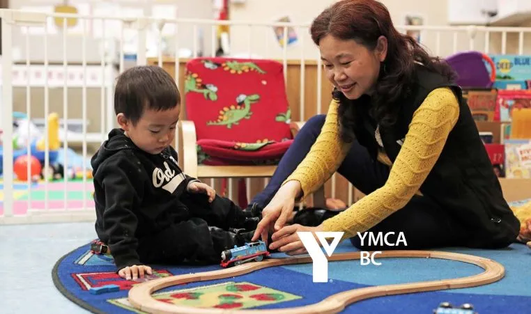 A toddler-aged boy and a YMCA child care staff team member play with a set of toy trains on the floor