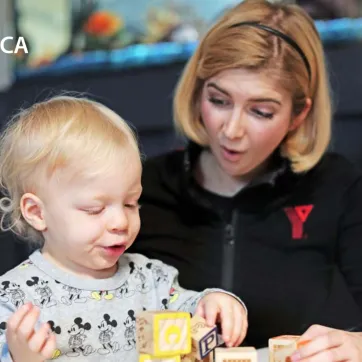 A YMCA child care staff team member sits with and reacts to a young child playing with blocks