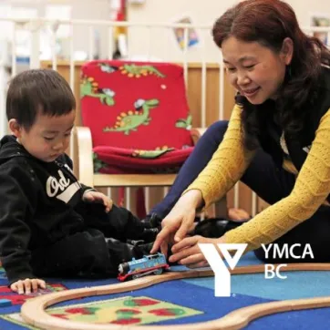 A toddler-aged boy and a YMCA child care staff team member play with a set of toy trains on the floor