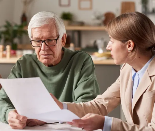 A woman working as a financial advisor reviews documents with an elderly man as they sit at a table together.