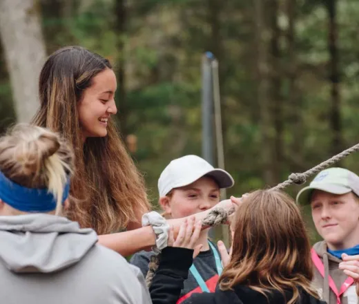 A female camp counsellor is holding a rope while surrounded by youth campers as they prepare for an activity.