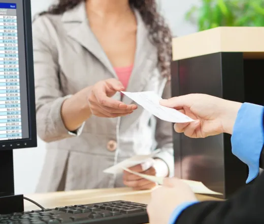 A woman hands a cheque over a desk to another woman sitting behind a computer with a spreadsheet open on the screen
