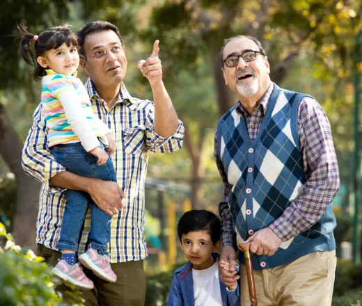 Two elderly gentlemen with a young boy and girl who appear to be their grandchildren. The man on the left is holding up the girl and pointing to something above them, the other man holds the boy's hand as they stand together and look up.
